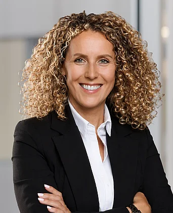 Porträt einer Person mit schulterlangem, lockigem Haar in Business-Kleidung. Die Person trägt ein schwarzes Sakko über einer weißen Bluse und steht mit verschränkten Armen. An einem Handgelenk ist eine Armbanduhr sichtbar. Der Hintergrund zeigt einen hellen, unscharfen Innenraum in Büroatmosphäre mit neutralen Farben und weicher Beleuchtung.