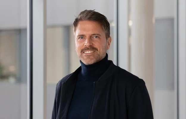 Porträt einer Person mit dunklem Rollkragenpullover und Jacke vor einer hellen Glasfassade.