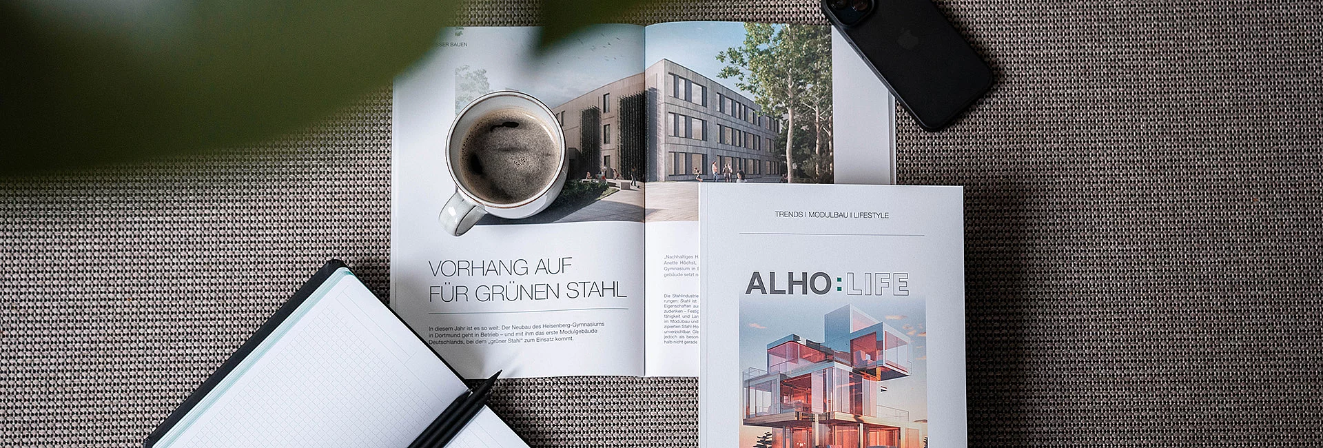 Eine aufgeschlagene Zeitschrift zeigt Architektur mit der Überschrift "VORHANG AUF FÜR GRÜNEN STAHL". Daneben liegt eine Tasse Kaffee auf einem Tisch, umgeben von Notizblock und Smartphone.