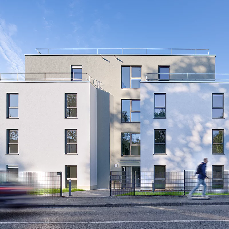 Ein modernes Wohngebäude mit großen Fenstern steht vor blauem Himmel. Im Vordergrund passiert ein Fußgänger; ein rotes Auto fährt vorbei.