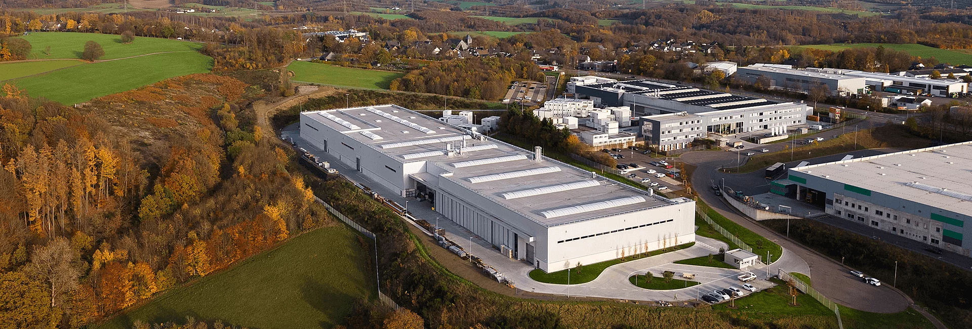 Aus der Vogelperspektive eine große weiße Industriehalle mit Laderampen und Parkplätzen, eingebettet in herbstliche Wälder, grüne Felder und angrenzende Gewerbegebäude.