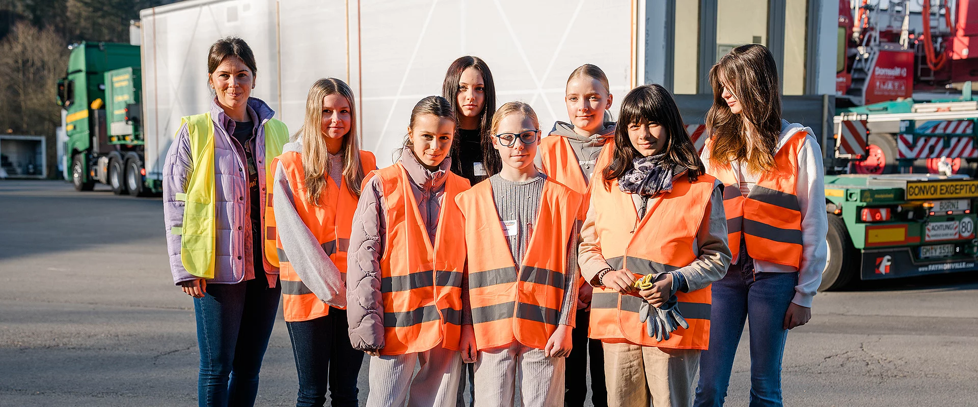 Eine Gruppe von Menschen trägt orangefarbene Warnwesten, steht auf einem Parkplatz vor Lastwagen. Es sind vorwiegend junge Frauen zu sehen, die in die Kamera schauen.