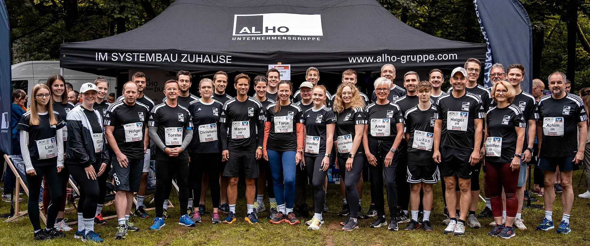 Eine Gruppe von Menschen in Sportkleidung steht vor einem Zelt mit der Aufschrift "ALHO Unternehmensgruppe". Sie tragen Namen und Nummern an ihren T-Shirts.