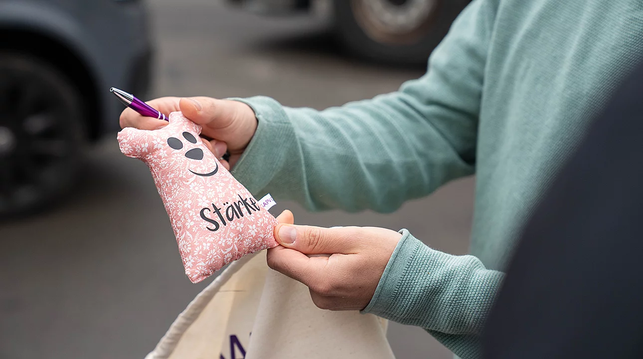 Person hält ein kleines rosafarbenes Stofftäschchen mit aufgedrucktem Smiley und dem Schriftzug „Stärke“ in der Hand, darin steckt ein Stift; Szene im Außenbereich.