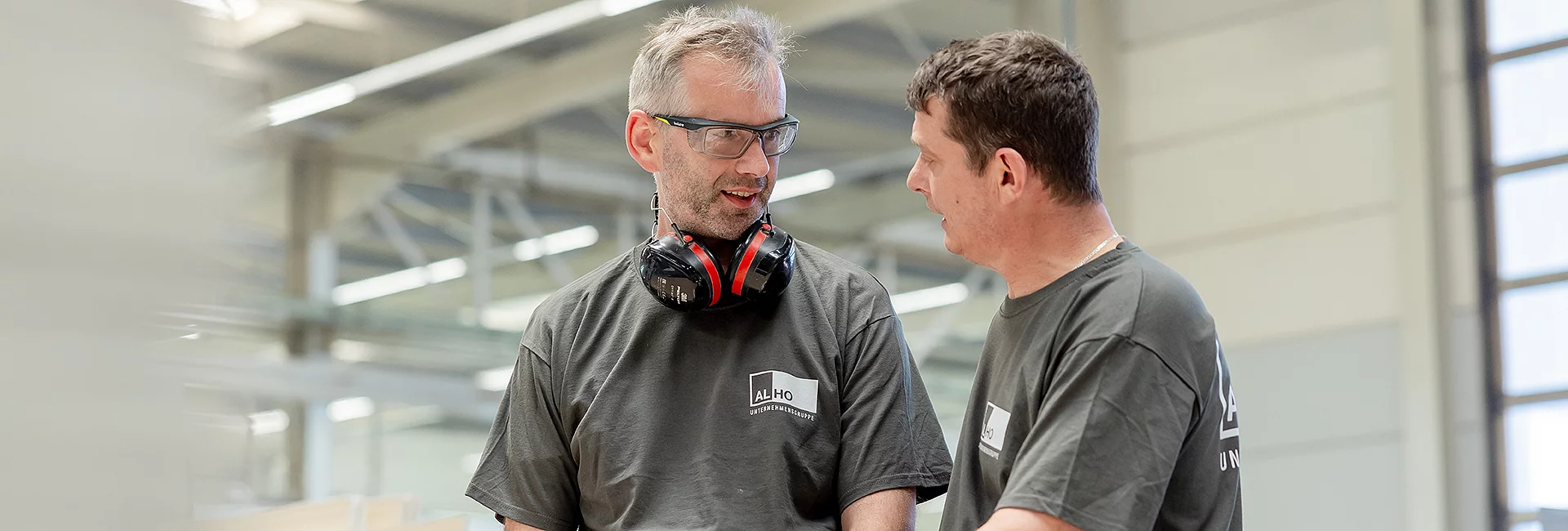 Zwei Männer tragen Arbeitskleidung mit "ALHO Unternehmensgruppe"-Logos, unterhalten sich in einer Fabrikumgebung. Einer trägt Schutzbrille und Gehörschutz um den Hals. Im Hintergrund sind Industrieanlagen erkennbar.