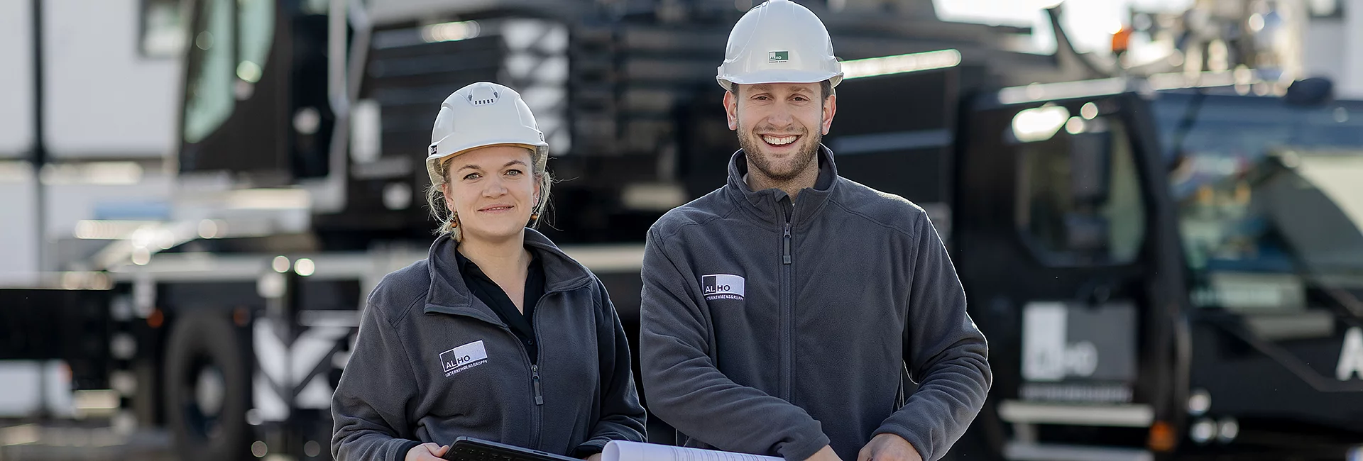 Zwei Personen in Arbeitskleidung und Helmen stehen nebeneinander. Beide lächeln, halten Pläne. Das ALHO-Logo ist auf ihrer Kleidung sichtbar. Im Hintergrund ist eine Baustelle.