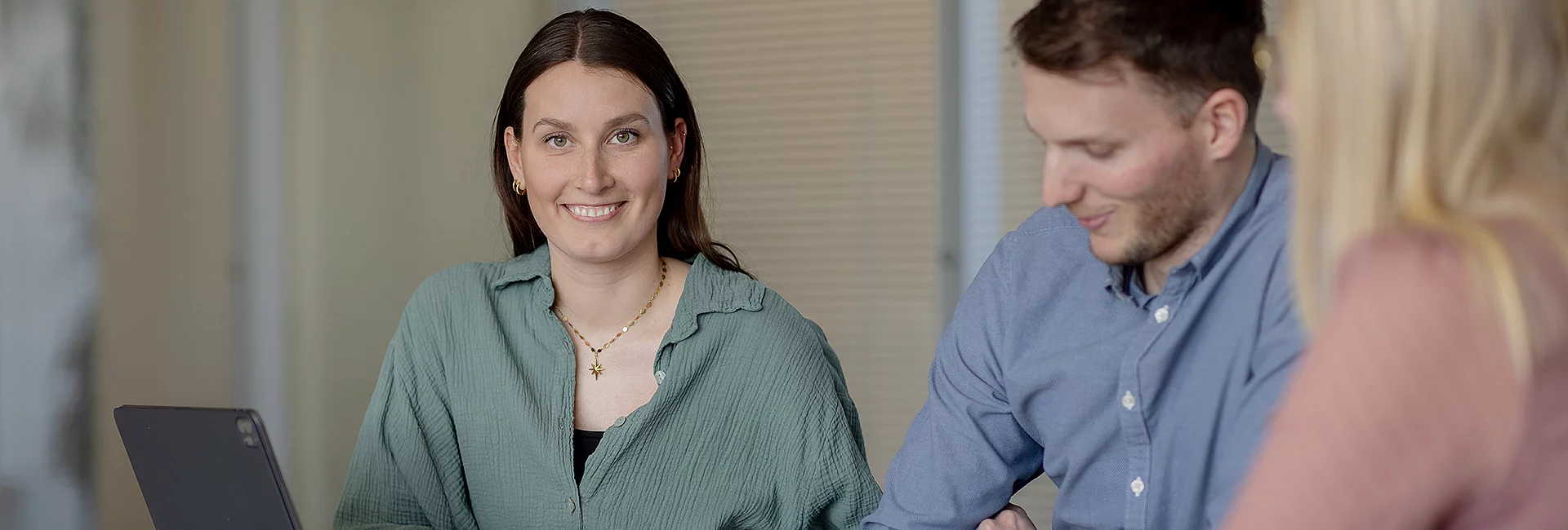 Eine lächelnde Frau sitzt vor einem Laptop und blickt in die Kamera, während zwei Kolleg:innen neben ihr arbeiten; heller Besprechungsraum mit geschlossenen Jalousien im Hintergrund.