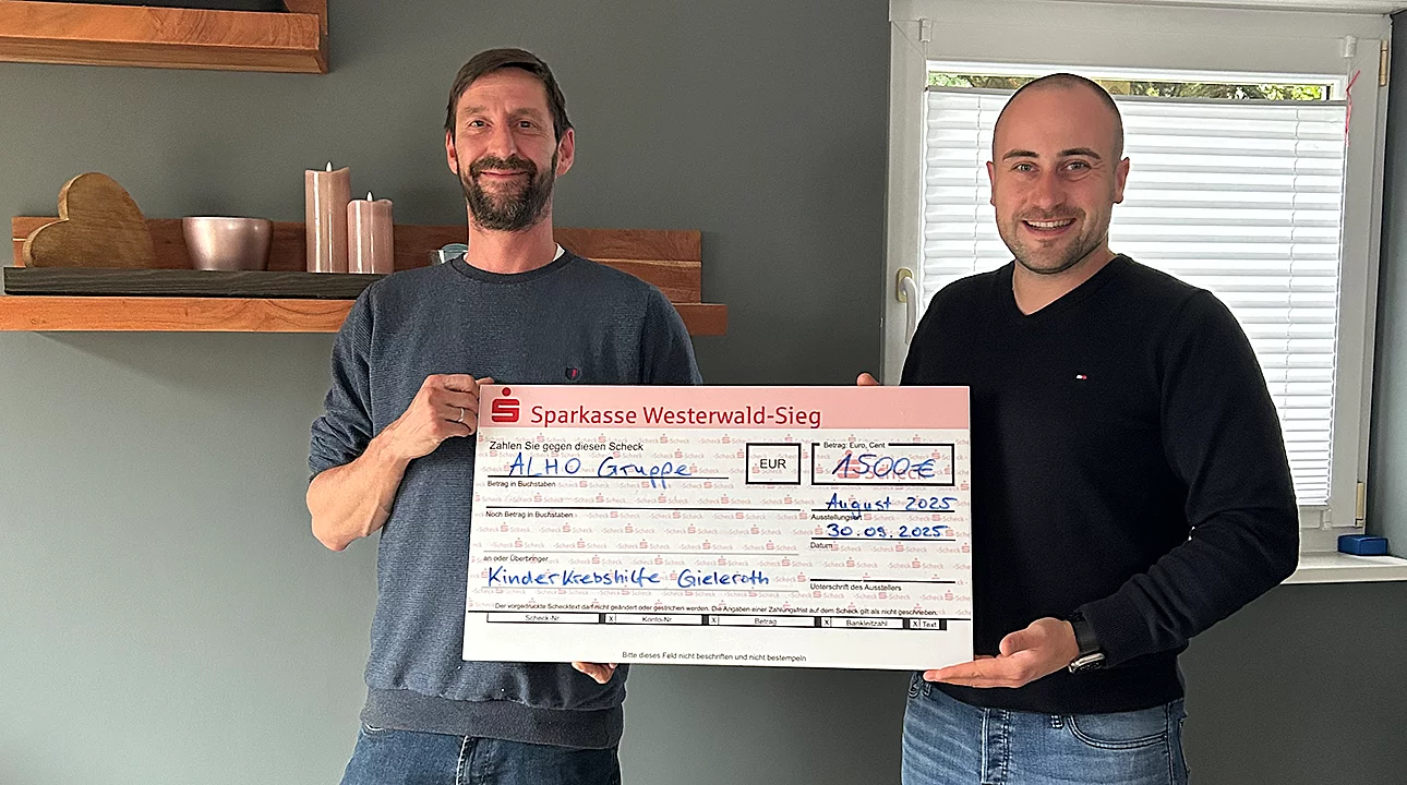 Zwei Männer halten einen großen Scheck der Sparkasse Westerwald-Sieg über 1.500 Euro. Text: „ALHO Gruppe, August 2025, Kindekrebshilfe Gieleroth, 30.05.2023.“ Sie stehen in einem Raum mit Regal im Hintergrund.