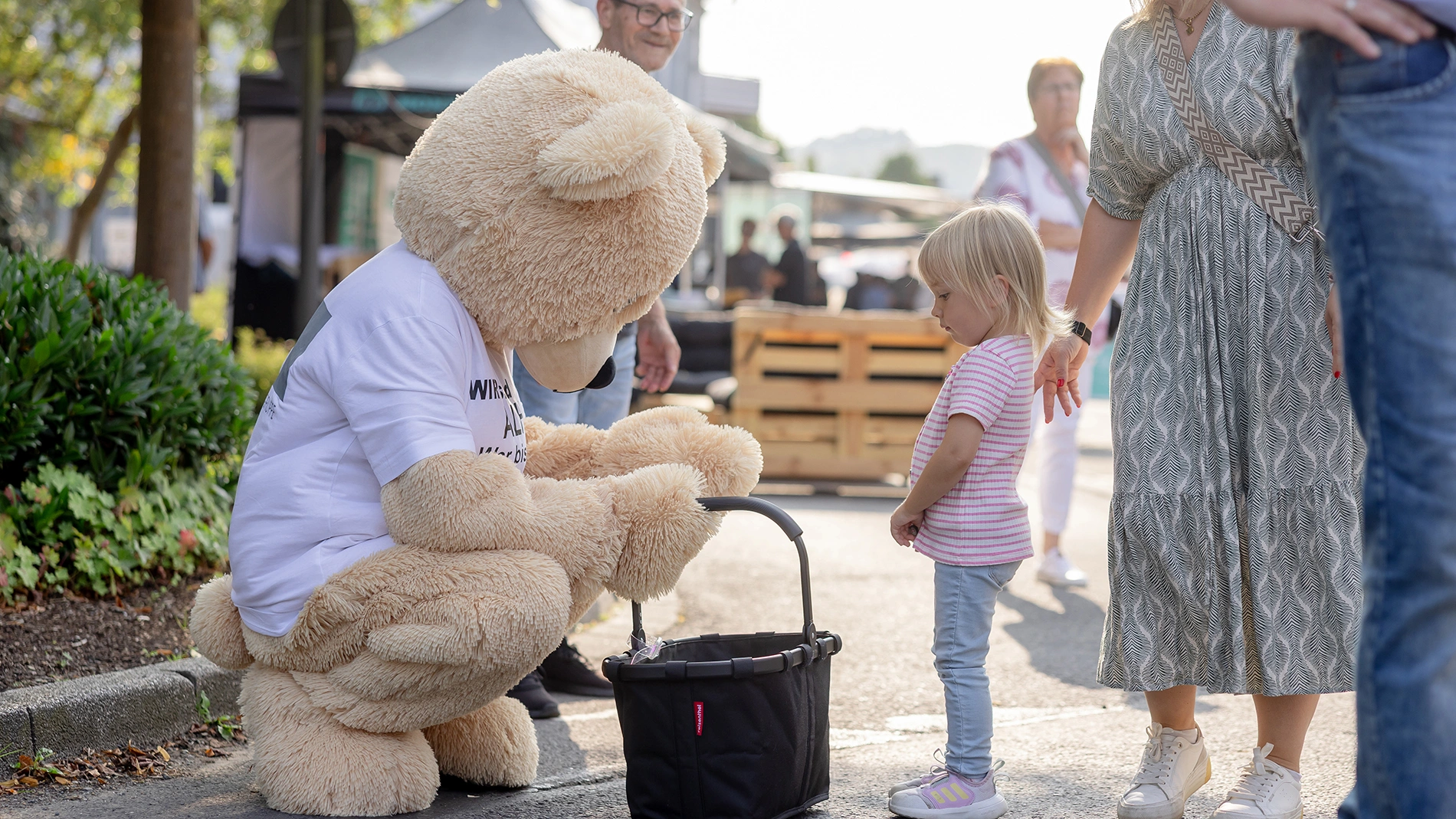 Ein Mensch im Plüschbärenkostüm kniet vor einem kleinen Kind auf der Straße, hält einen Korb. Umgeben von Passanten in sommerlicher Kleidung. Der Bär trägt ein weißes T-Shirt mit unlesbarem Text.
