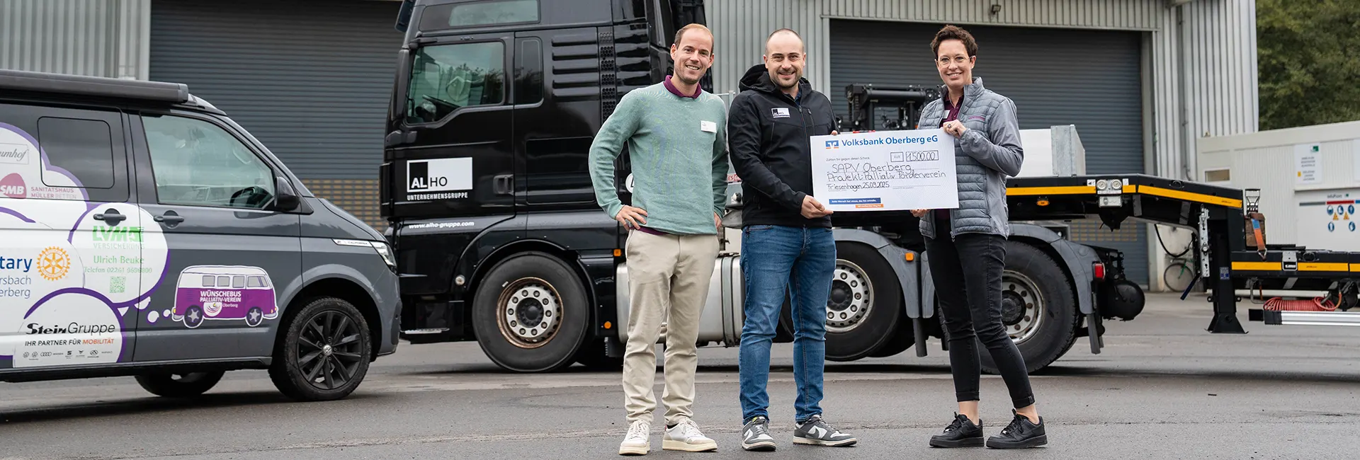 Drei Personen halten einen großen Scheck mit der Aufschrift „Volksbank Oberberg eG 1500,00 € SAPV Oberberg“, vor einem schwarzen LKW und einem grauen Lieferwagen auf einem Industriegelände.