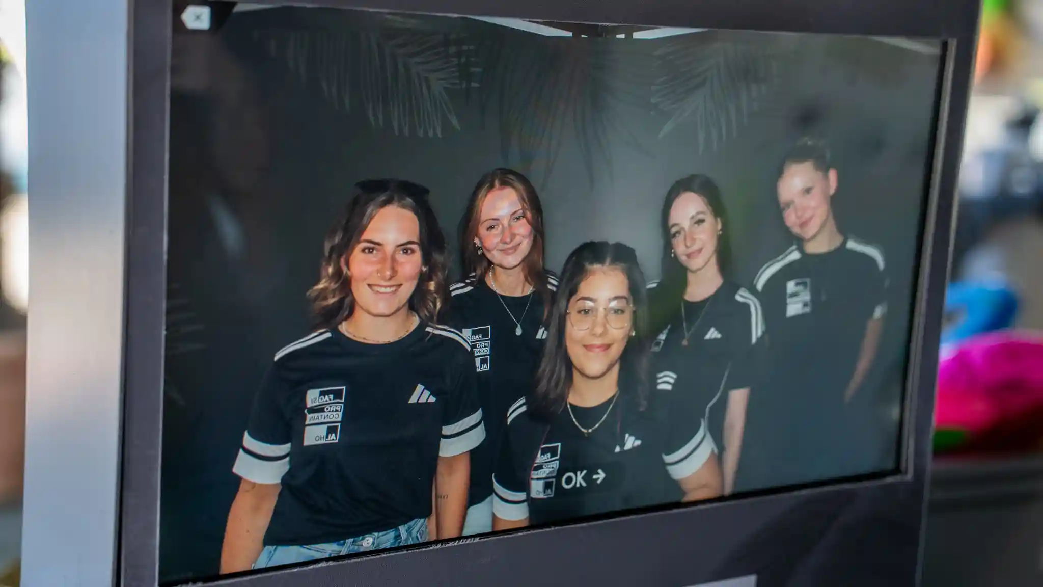 Auf einem Bildschirm ist ein Gruppenfoto von fünf Personen in schwarzen Trikots mit weißen Streifen zu sehen. Die Aufnahme wurde in einer Fotobox oder an einer digitalen Fotostation erstellt.