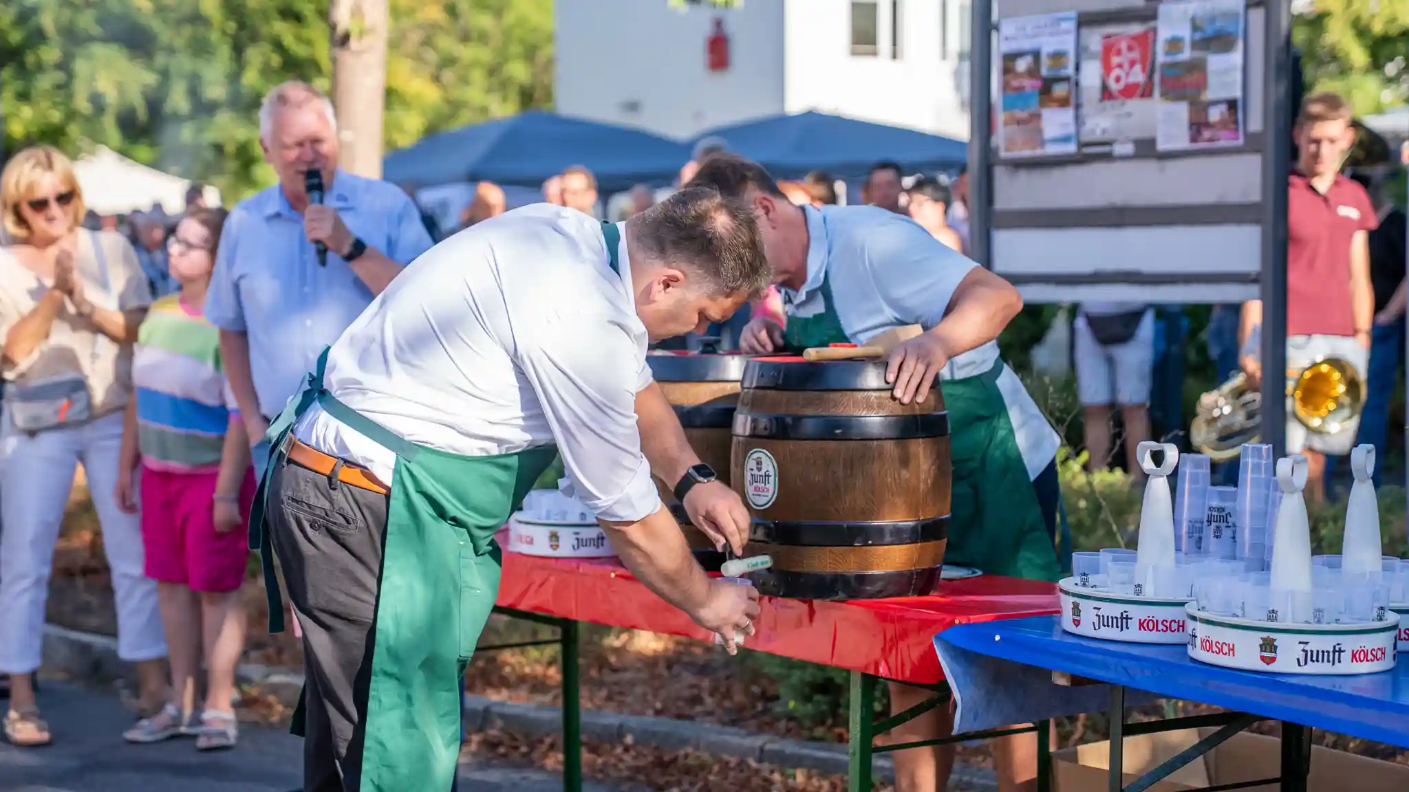 Zwei Personen mit grünen Schürzen zapfen an einem Holzfass Getränke aus. Im Hintergrund stehen Besucherinnen und Besucher einer Veranstaltung sowie eine Person mit Mikrofon.