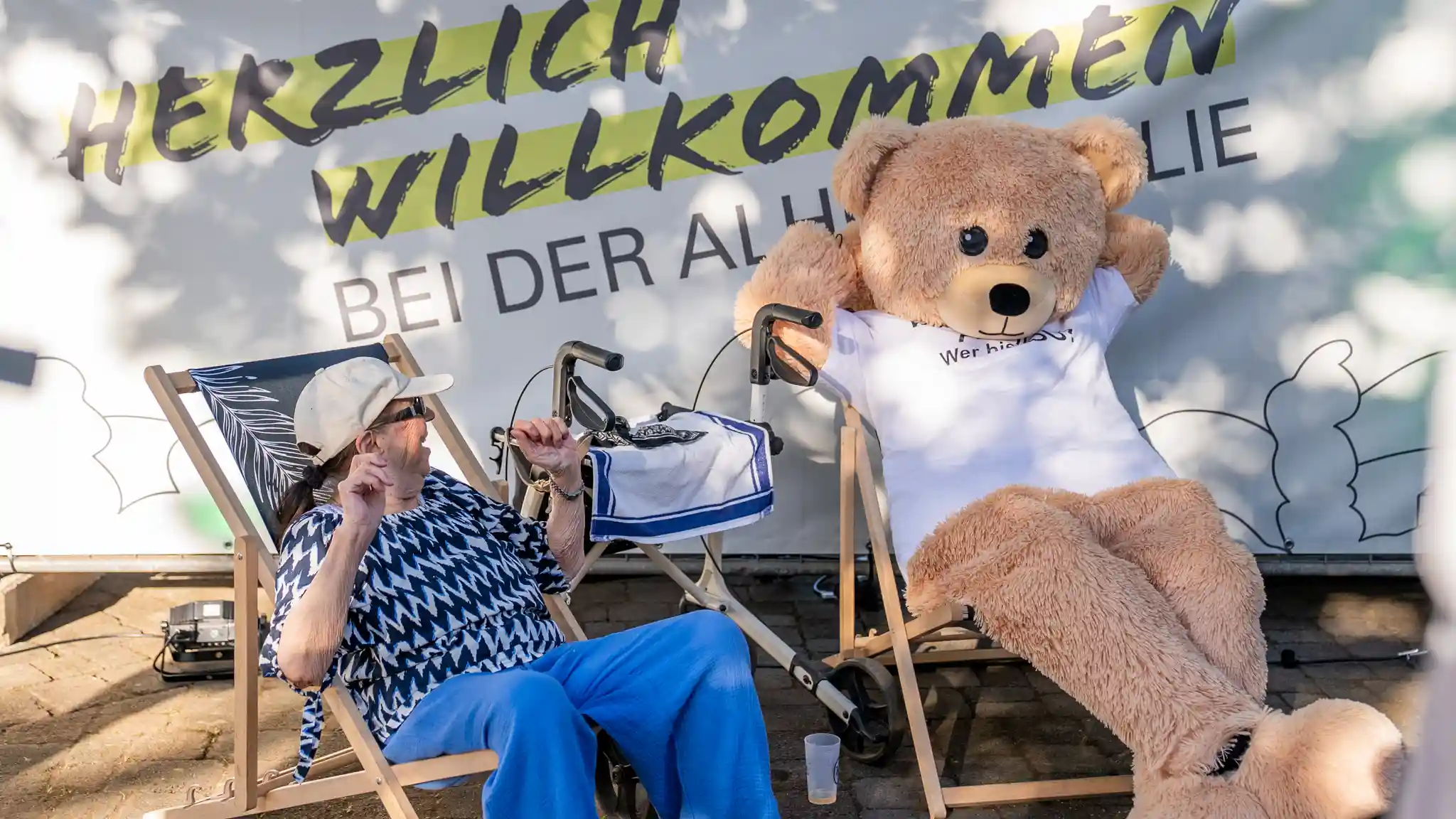 Ein großes Bärenmaskottchen sitzt in einem Liegestuhl neben einer Person vor einer Wand mit der Aufschrift „HERZLICH WILLKOMMEN BEI DER ALHO FAMILIE“. Zwischen den Liegestühlen steht eine Gehhilfe.