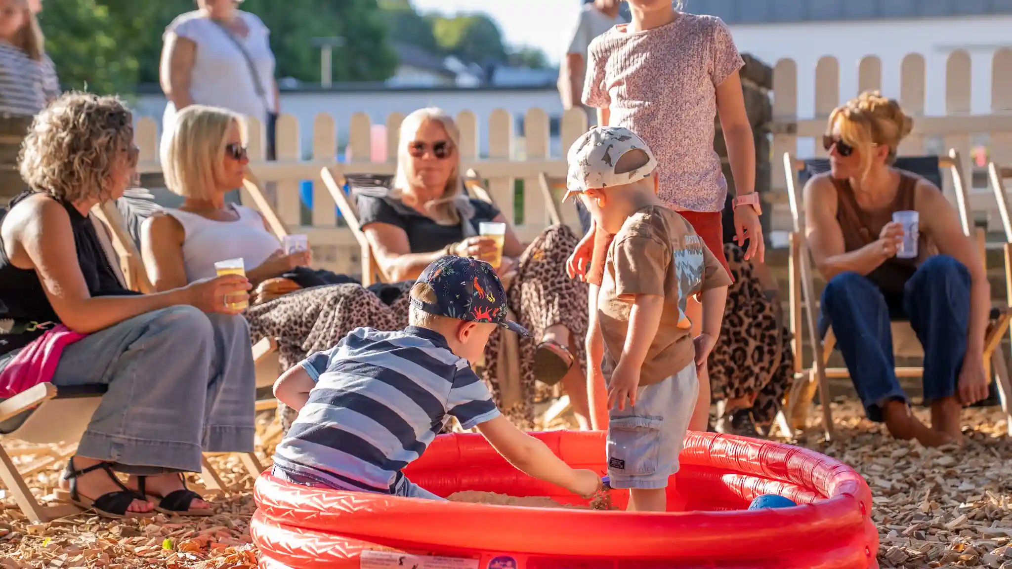 Kinder spielen in einem roten Sandkasten, während mehrere Personen auf Liegestühlen sitzen und sich auf einer Veranstaltung im Freien unterhalten. Ein heller Zaun umrahmt den Bereich.